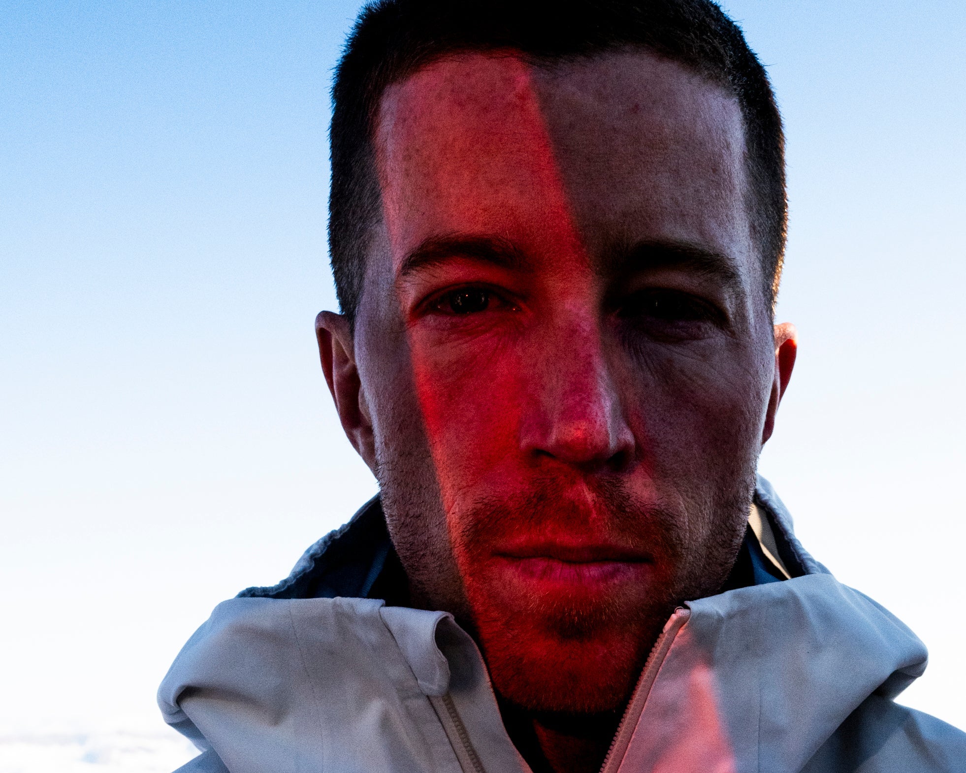 Close up of Shaun White's face standing outside with a red glare over one eye and down to the opposite shoulder with a blue sky background.