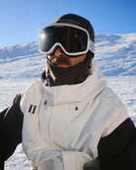 A close-up shot of a professional snowboarder wearing the WHITESPACE Dual Tone Goggle (Cream) and WHITESPACE Alpine X Jacket (White) set against a snowy mountain background under a clear blue sky