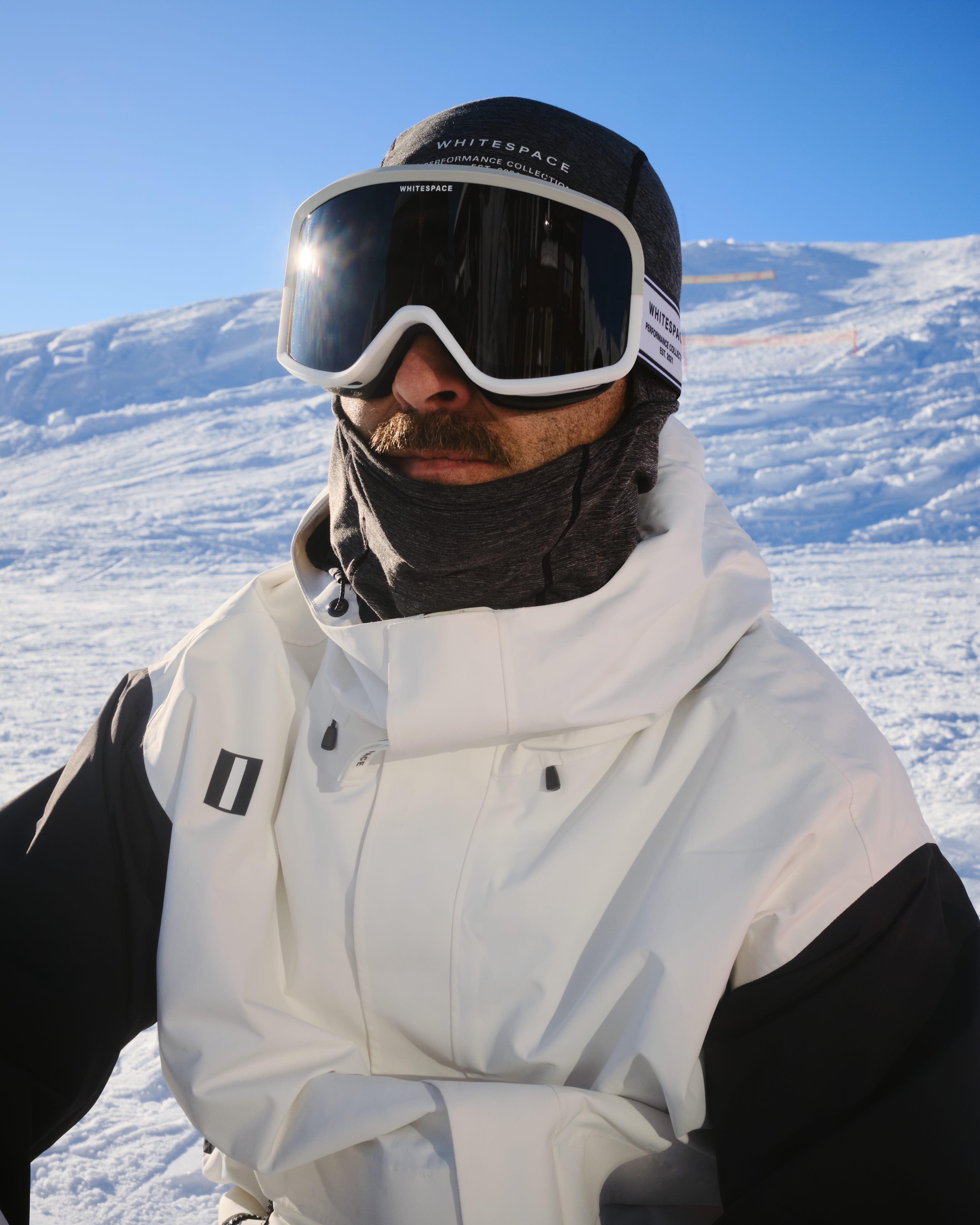 A close-up shot of a professional snowboarder wearing the WHITESPACE Dual Tone Goggle (Cream) and WHITESPACE Alpine X Jacket (White) set against a snowy mountain background under a clear blue sky