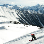 Person snowboarding down a mountain side with a beautiful background of snow, mountains peaks, and trees.