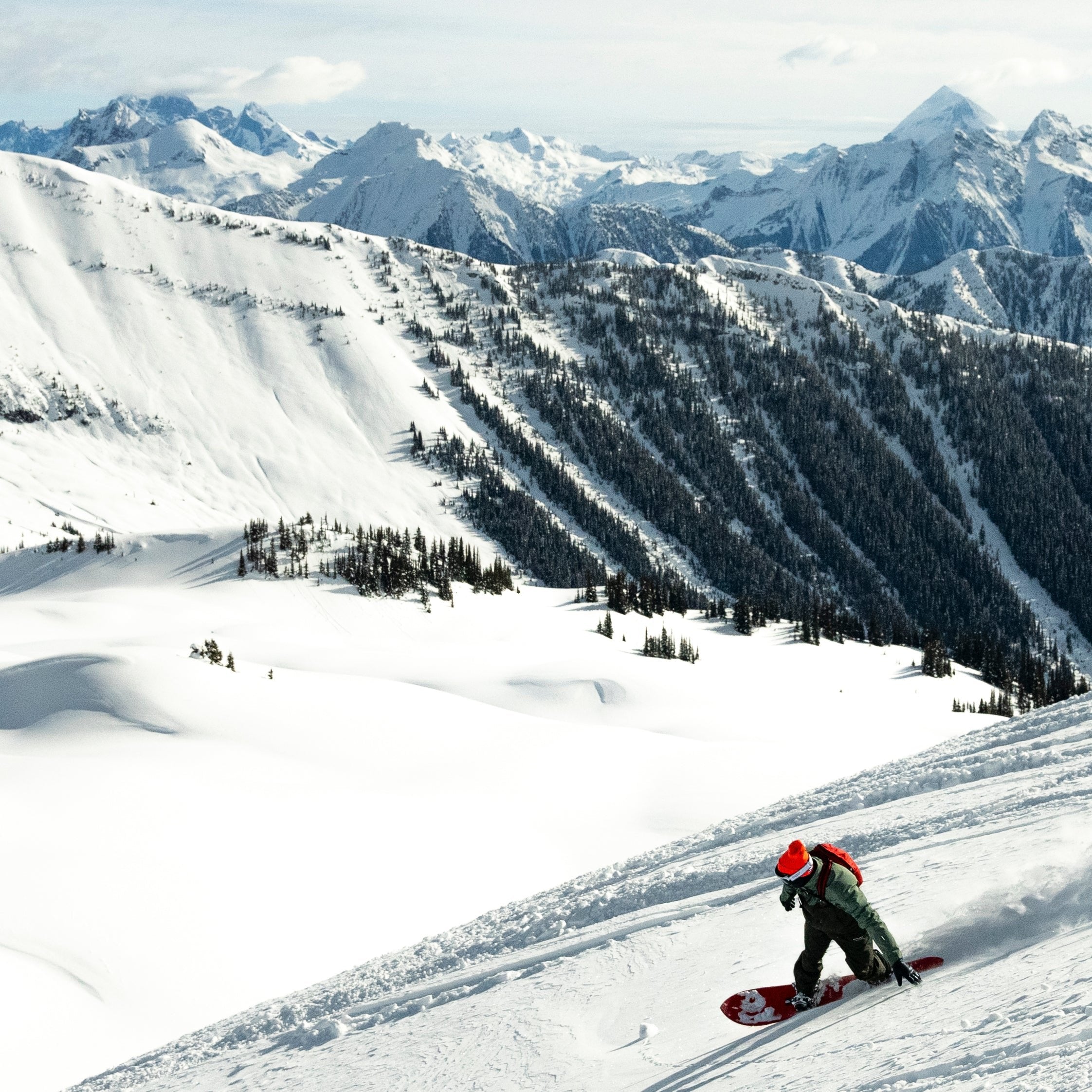 Person snowboarding down a mountain side with a beautiful background of snow, mountains peaks, and trees.