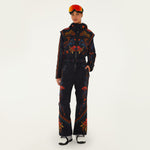 Model wearing Farm Rio Black Macaw jumpsuit, a black jumpsuit with orange and blue floral designs, orange ski goggles, and white shoes, on a white background.