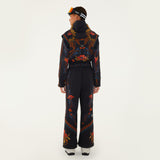 Model, from the back, wearing Farm Rio Black Macaw jumpsuit, a black jumpsuit with orange and blue floral designs, black beanie, black and orange ski goggles, and white shoes, on a white background.