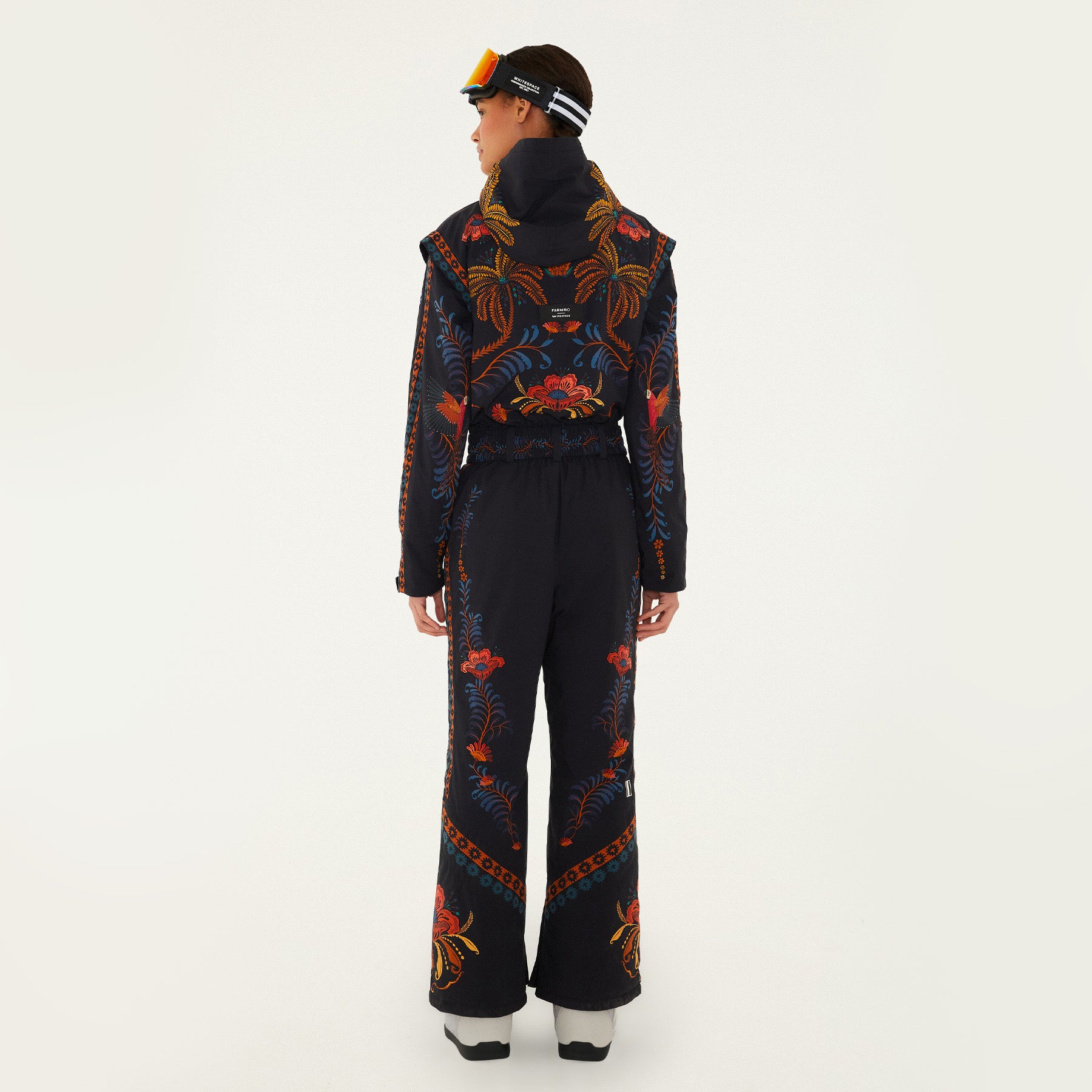 Model, from the back, wearing Farm Rio Black Macaw jumpsuit, a black jumpsuit with orange and blue floral designs, black beanie, black and orange ski goggles, and white shoes, on a white background.