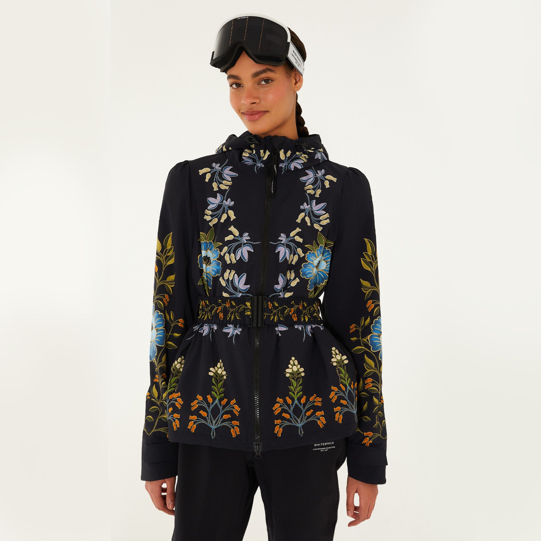 Model wearing Farm Rio Garden belted jacket, a black jacket with blue, orange and white flower design, black ski goggles, and black pants, on a white background.