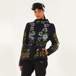 Model wearing Farm Rio Garden belted jacket, a black jacket with blue, orange and white flower design, black ski goggles, and black pants, on a white background.
