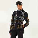 Model wearing Farm Rio Garden belted jacket, a black jacket with blue, orange and white flower design, black ski goggles, and black pants, on a white background.