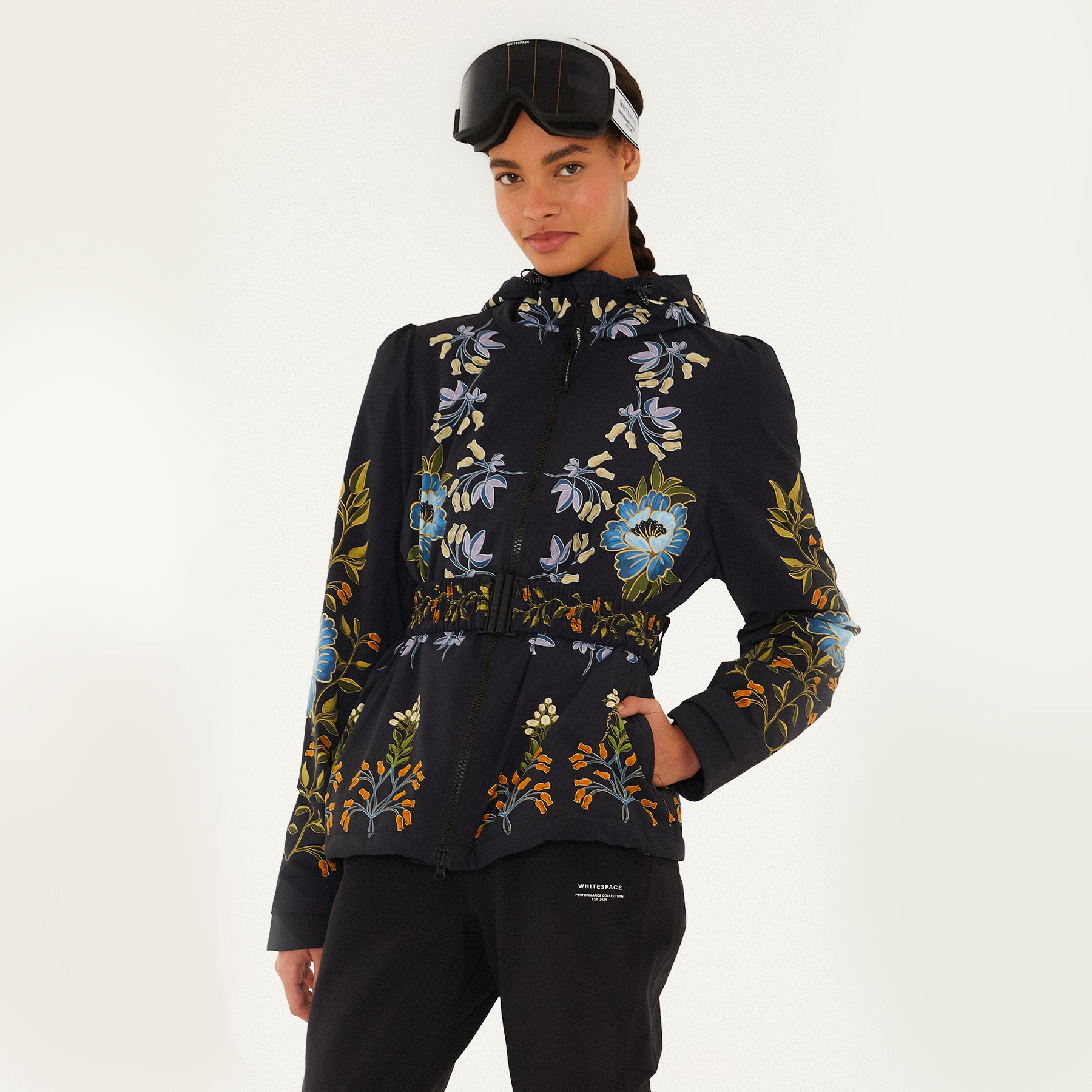 Model wearing Farm Rio Garden belted jacket, a black jacket with blue, orange and white flower design, black ski goggles, and black pants, on a white background.