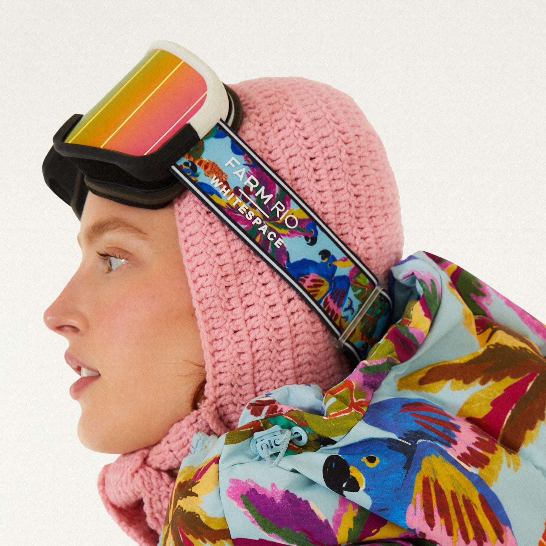 Head shot of model looking left, wearing Farm Rio/WHITESPACE ski googles over a crochet hood and light blue puffer jacket with macaw graphics, on a white background.