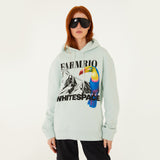 Model wearing Farm Rio/ WHITESPACE light blue hoodie with toucan and mountain graphic design, dark sunglasses, and black pants.