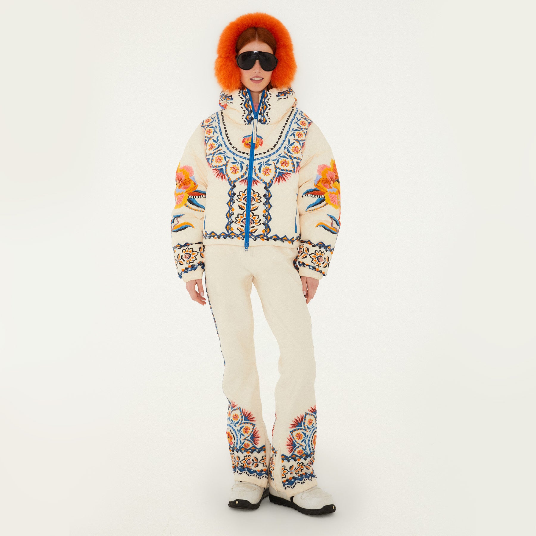 Model wearing Farm Rio Lucy Floral Puffer Jacket and bibs, cream colored jacket and bib with blue and yellow floral designs, white shoes, dark sunglasses, and fluffy orange ear muffs, on a white background
