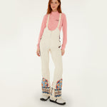 Model wearing Farm Rio Lucy floral riding bib pants, cream colored bibs with orange and blue floral designs around the bottom of the legs and up the sides to the waist over a pink shirt, and white shoes, on a white background.