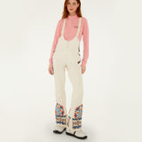 Model wearing Farm Rio Lucy floral riding bib pants, cream colored bibs with orange and blue floral designs around the bottom of the legs and up the sides to the waist over a pink shirt, and white shoes, on a white background.