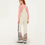 Model, from the back, wearing Farm Rio Lucy floral riding bib pants, cream colored bibs with orange and blue floral designs around the bottom of the legs and up the sides to the waist over a pink shirt, and white shoes, on a white background.
