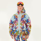 Model wearing Farm Rio Macaw puffer jacket, a light blue jacket with macaw graphic design, matching pants, pink and orange ski goggles, and a crochet hood, on a white background.