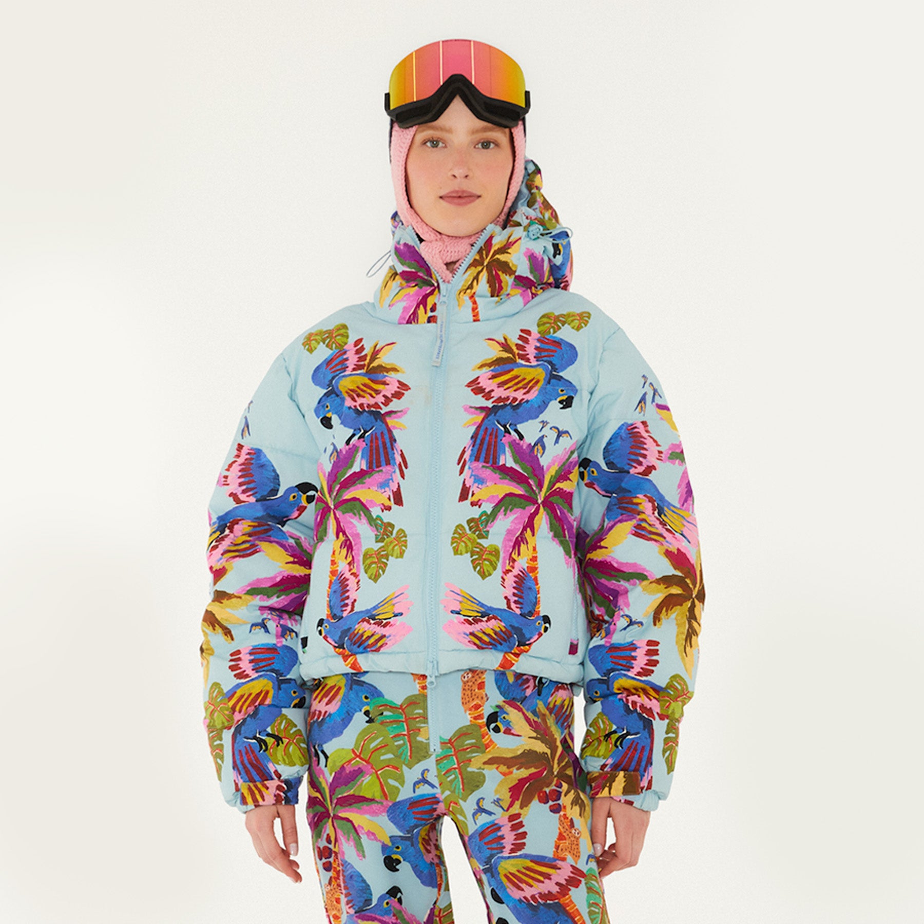 Model wearing Farm Rio Macaw puffer jacket, a light blue jacket with macaw graphic design, matching pants, pink and orange ski goggles, and a crochet hood, on a white background.