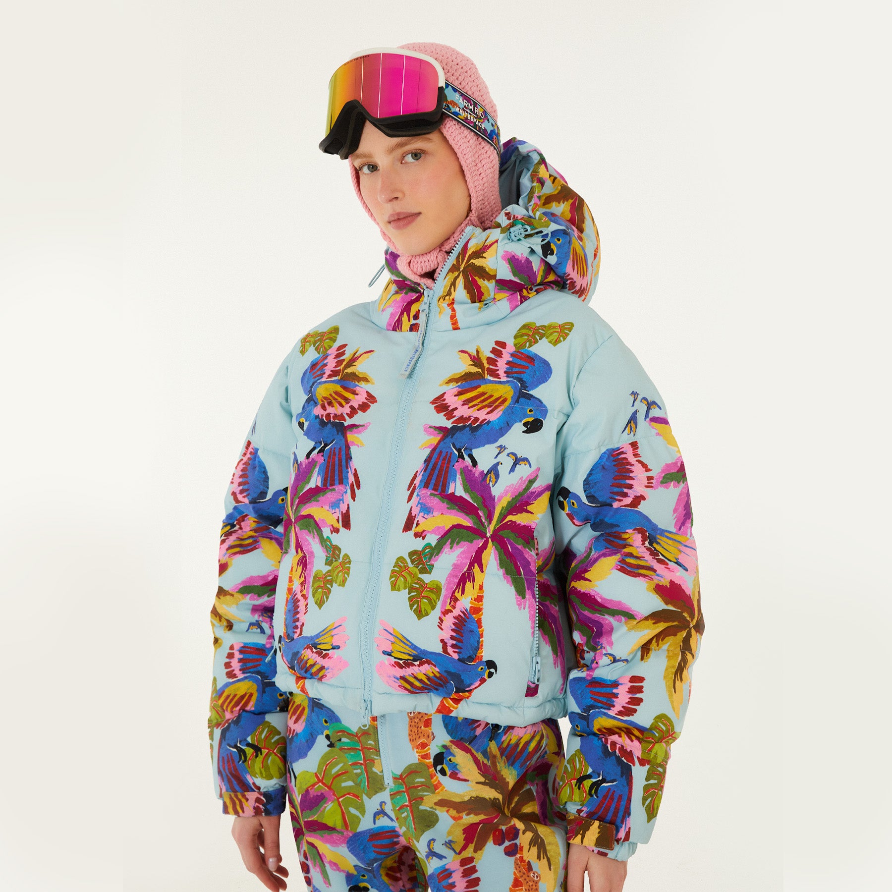 Model wearing Farm Rio Macaw puffer jacket, a light blue jacket with macaw graphic design, matching pants, pink and orange ski goggles, and a crochet hood, on a white background.