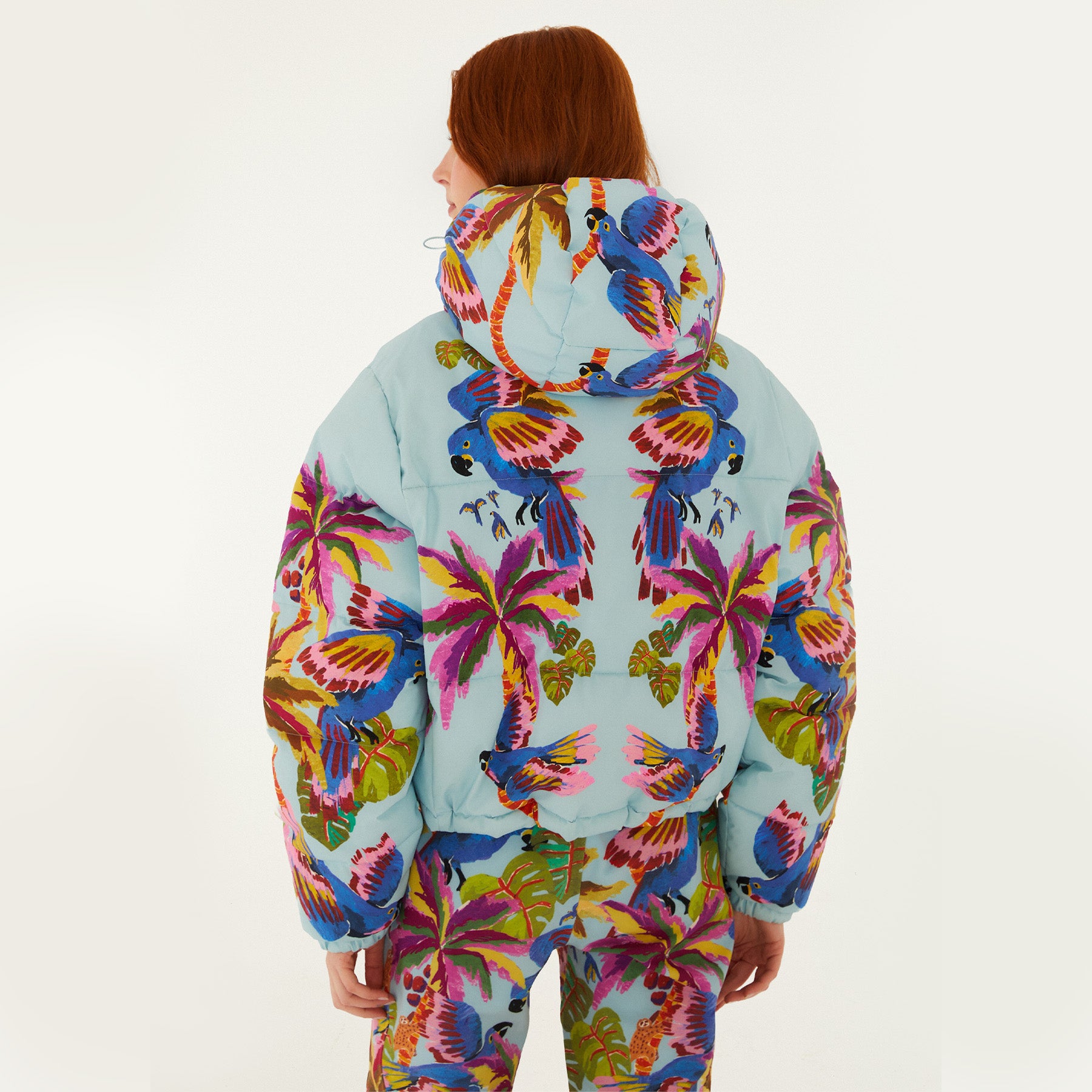 Model, from the back, wearing Farm Rio Macaw puffer jacket, a light blue jacket with macaw graphic design and matching pants, on a white background.