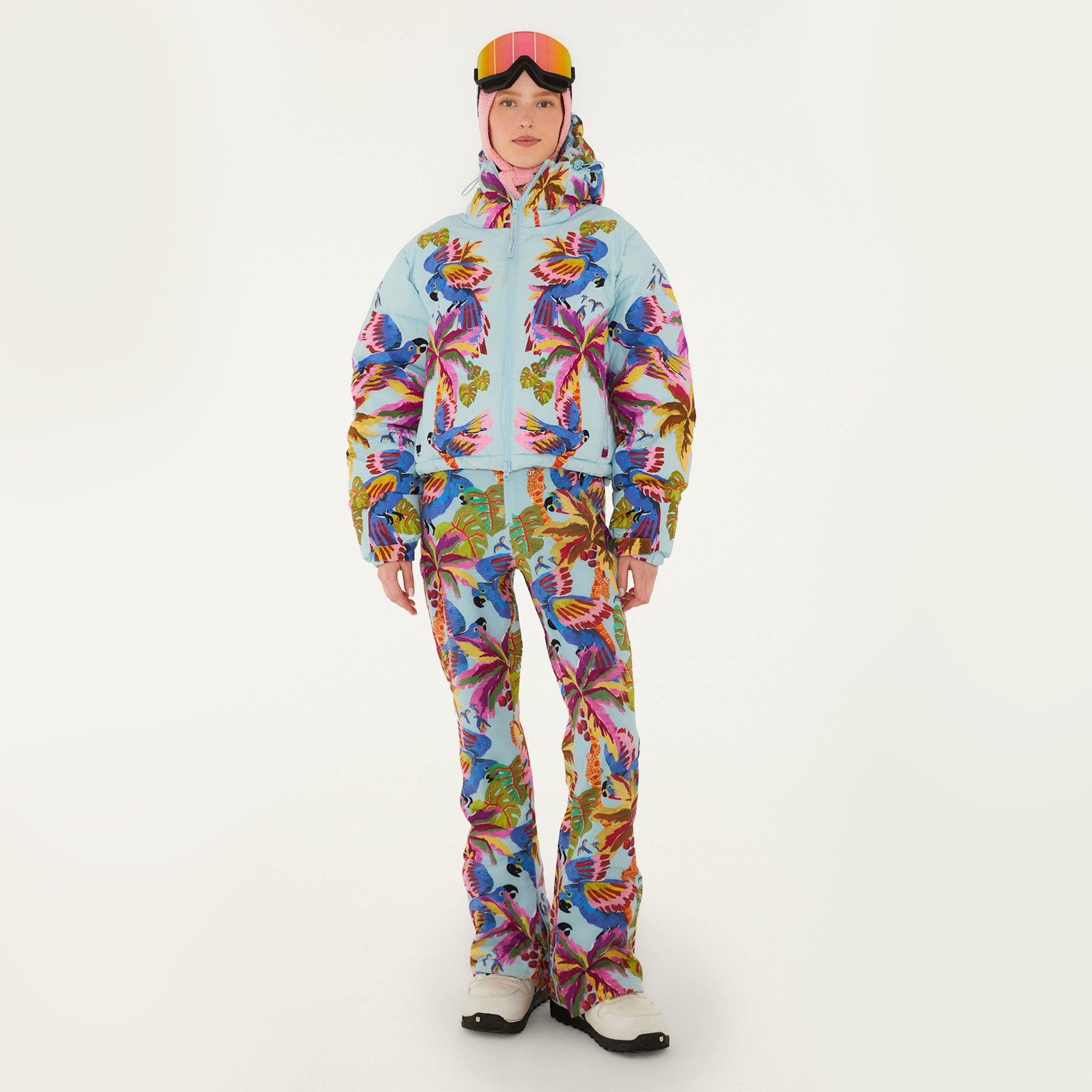 Model wearing Farm Rio Macaw riding bib and puffer jacket, a light blue bib and jacket with macaw graphic design, matching pants, pink and orange ski goggles, a crochet hood, and white shoes, on a white background.