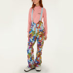 Model wearing Farm Rio Macaw riding bib pants, light blue colored bibs with blue macaws and tropical leaves over the entire pants, a peach long sleeved shirt, and white shoes, on a white background.