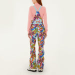 Model, from the back, wearing Farm Rio Macaw riding bib pants, light blue colored bibs with blue macaws and tropical leaves over the entire pants, a peach long sleeved shirt, and white shoes, on a white background.