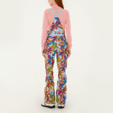 Model, from the back, wearing Farm Rio Macaw riding bib pants, light blue colored bibs with blue macaws and tropical leaves over the entire pants, a peach long sleeved shirt, and white shoes, on a white background.