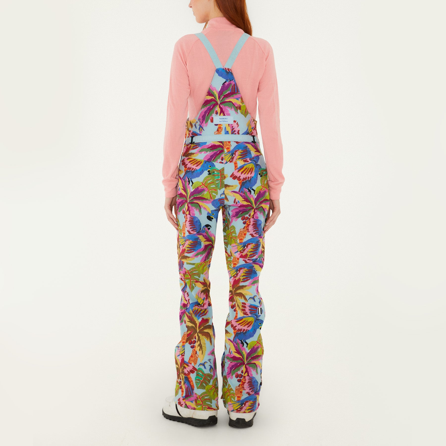 Model, from the back, wearing Farm Rio Macaw riding bib pants, light blue colored bibs with blue macaws and tropical leaves over the entire pants, a peach long sleeved shirt, and white shoes, on a white background.