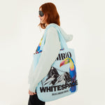 Model, from the side, facing left, with the Farm Rio/WHITESPACE puffer bag, a light blue bag with toucan and mountain graphics, over the shoulder, wearing a light blue hoodie, black pants, and dark sunglasses, on a white background.