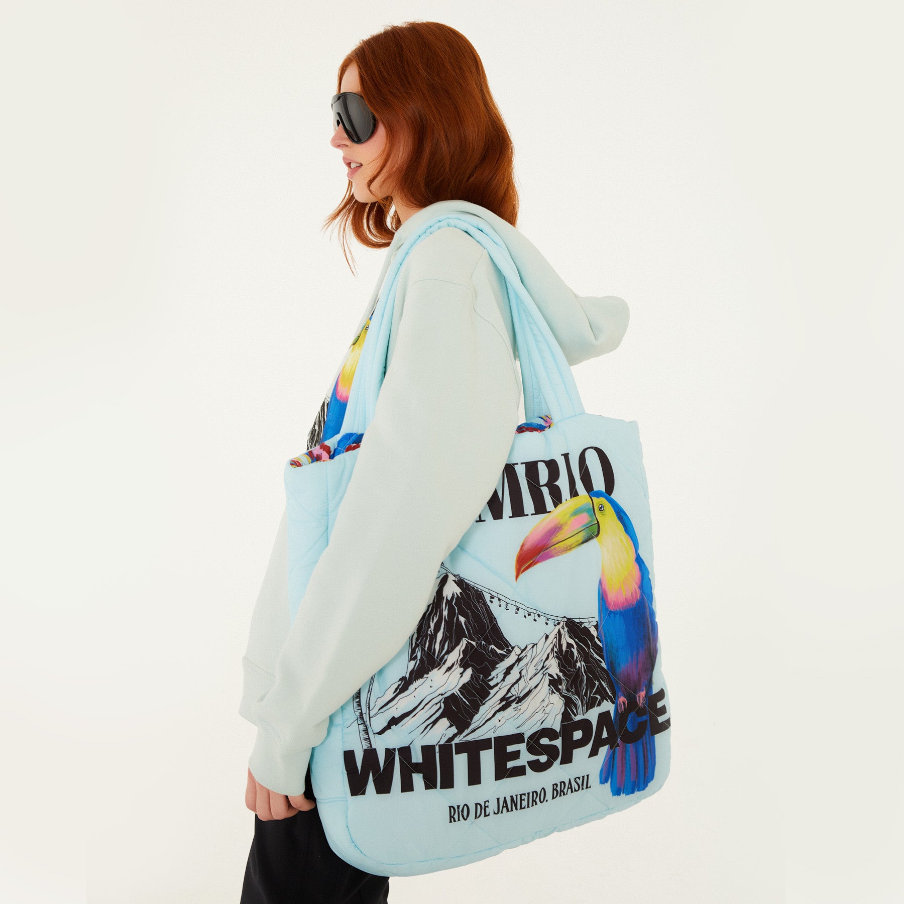Model, from the side, facing left, with the Farm Rio/WHITESPACE puffer bag, a light blue bag with toucan and mountain graphics, over the shoulder, wearing a light blue hoodie, black pants, and dark sunglasses, on a white background.