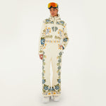 Model wearing Farm Rio Winter Garden jumpsuit, a cream jumpsuit with green and yellow floral designs, white shoes, a beige beanie and orange ski goggles, on a white background.