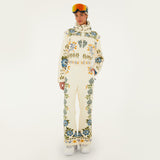 Model wearing Farm Rio Winter Garden jumpsuit, a cream jumpsuit with green and yellow floral designs, white shoes, a beige beanie and orange ski goggles, on a white background.