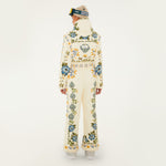 Model, from the back, wearing Farm Rio Winter Garden jumpsuit, a cream jumpsuit with green and yellow floral designs, white shoes, a beige beanie and ski goggles, on a white background.