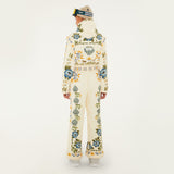 Model, from the back, wearing Farm Rio Winter Garden jumpsuit, a cream jumpsuit with green and yellow floral designs, white shoes, a beige beanie and ski goggles, on a white background.