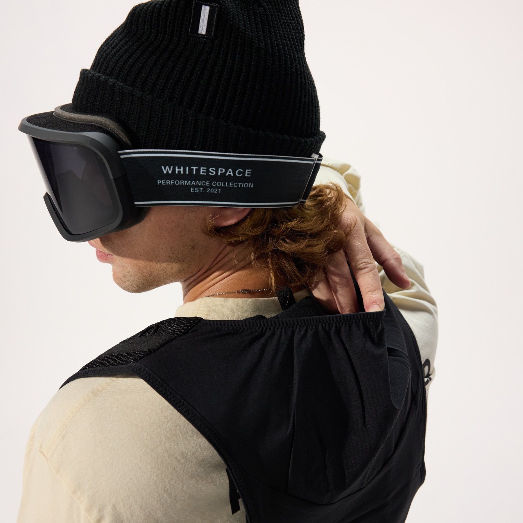 Model wearing black ski goggles and beanie, showing the behind the neck storage pocket on the black cargo vest.