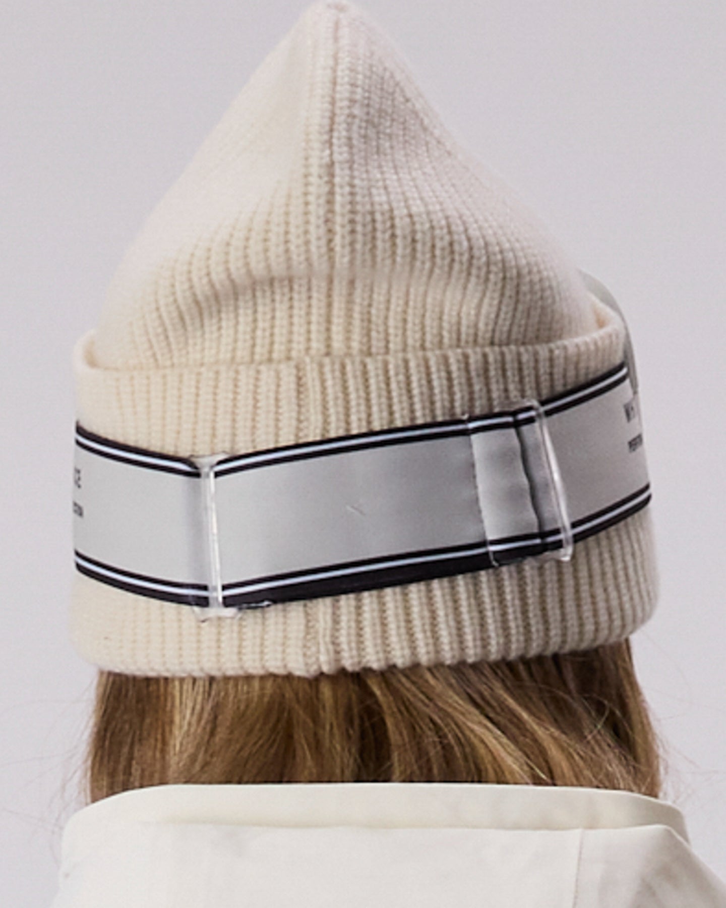 A close-up of a person wearing a cream-colored ribbed knit beanie with a wide white elastic band featuring black stripes and clear plastic sliders, viewed from the back.  This shows the WHITESPACE Dual Tone goggles in cream from the back.