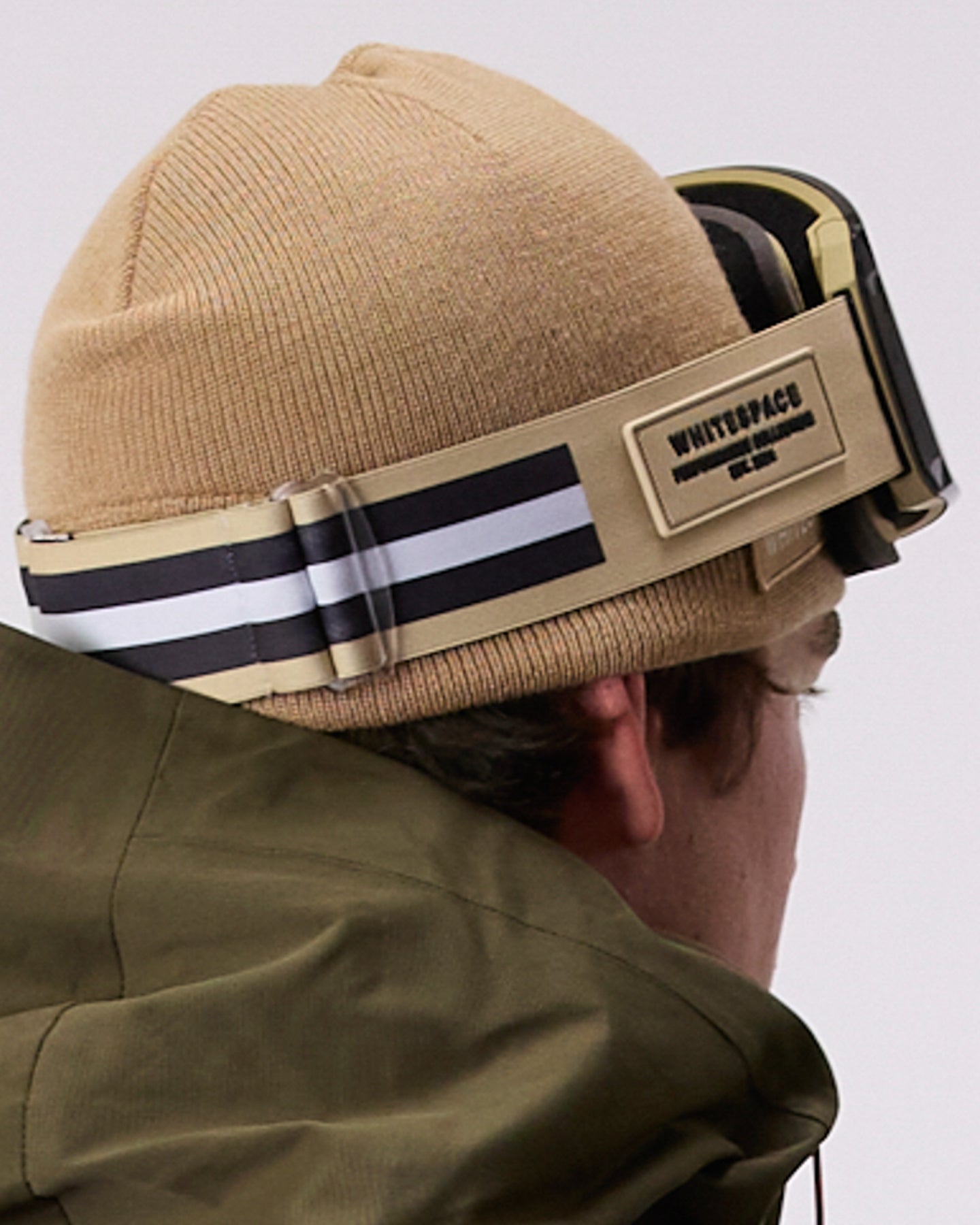 A close-up side profile of a person wearing a tan beanie, WHITESPACE MD Icon goggles with a tan strap, and a green jacket against a light gray background.