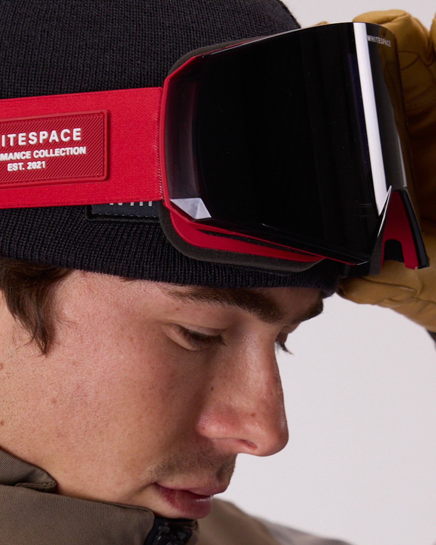 Close-up of a person wearing WHITESPACE MD Icon goggles in red, black ski goggles with a red frame featuring a red patch that reads 'WHITESPACE PERFORMANCE COLLECTION EST. 2021', over a black beanie