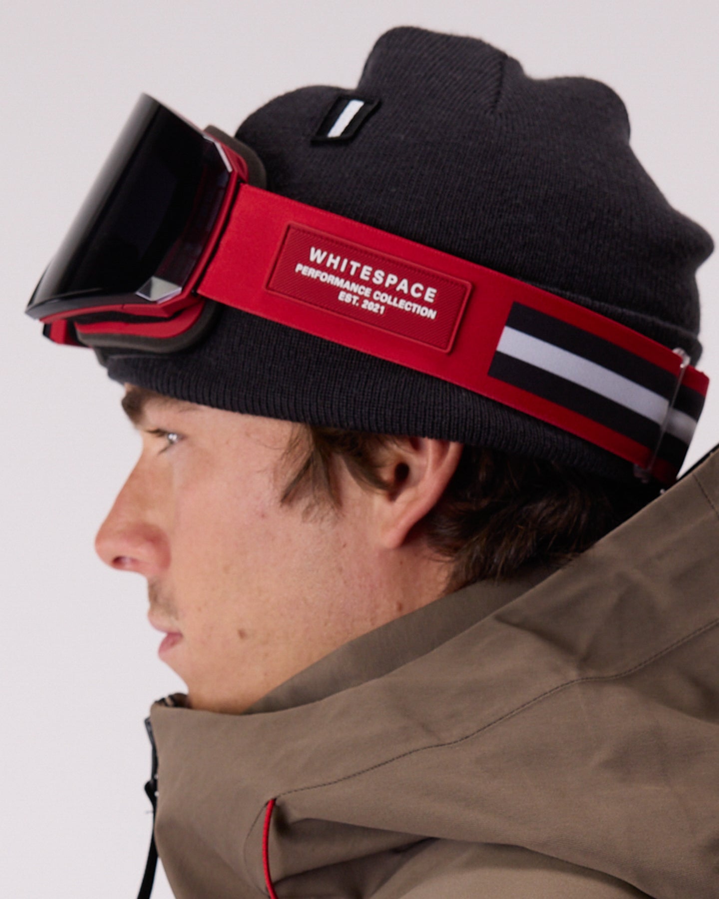 A close-up side profile of a person wearing a black beanie, WHITESPACE MD Icon goggles with a red strap, and a brown jacket against a light gray background. The goggle strap features a prominent red panel with the branding 'WHITESPACE PERFORMANCE COLLECTION'