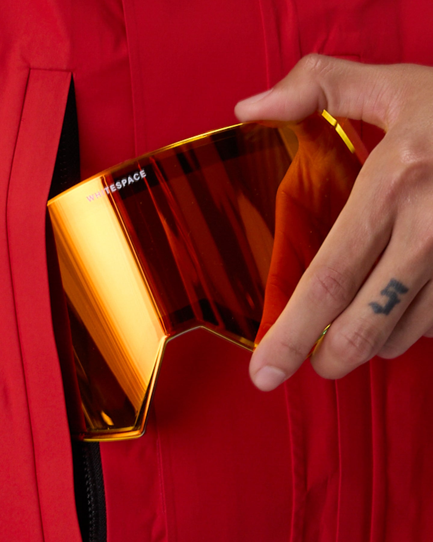 A close-up of a hand sliding a large, curved shield lens, into a red zippered pocket of a red jacket. The lens has a reflective gradient of orange, yellow, and red.  This is the alternate magnetic lens that comes with the WHITESPACE MD Icon Goggle in red.
