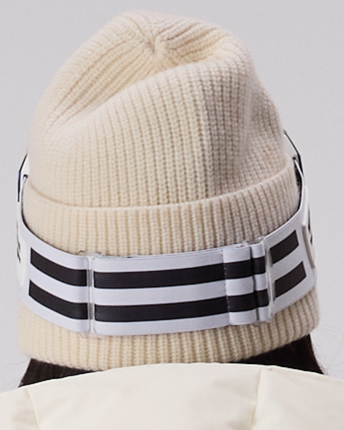 A close-up of a cream-colored ribbed knit beanie hat with a wide white elastic band featuring three black stripes.  This shows the WHITESPACE MD Icon goggles in white from the back.