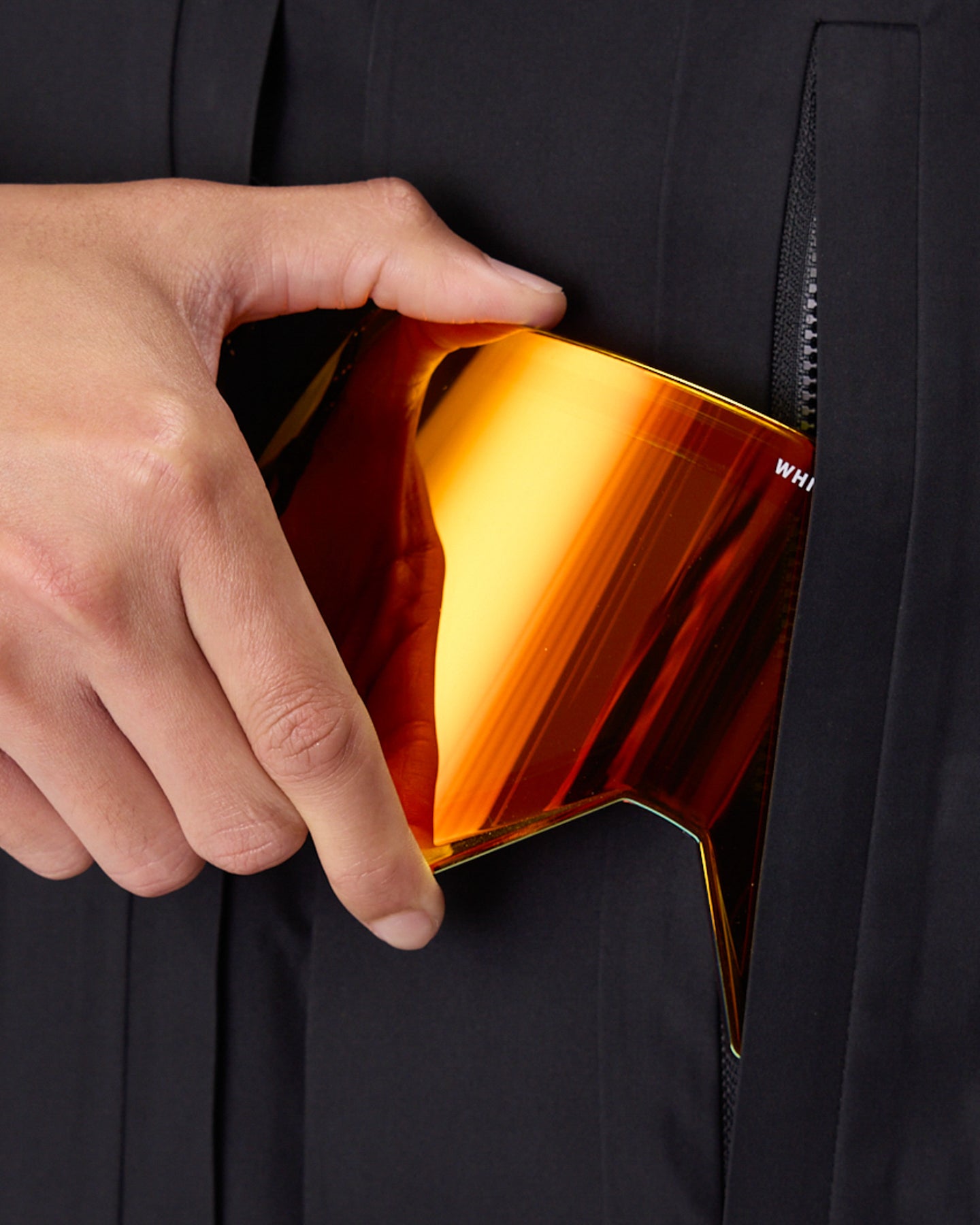 A close-up of a hand sliding a large, curved shield lens, into a black zippered pocket of a dark jacket. The lens has a reflective gradient of orange, yellow, and red.  This is the alternate magnetic lens that comes with the WHITESPACE MD Icon Goggle in white.