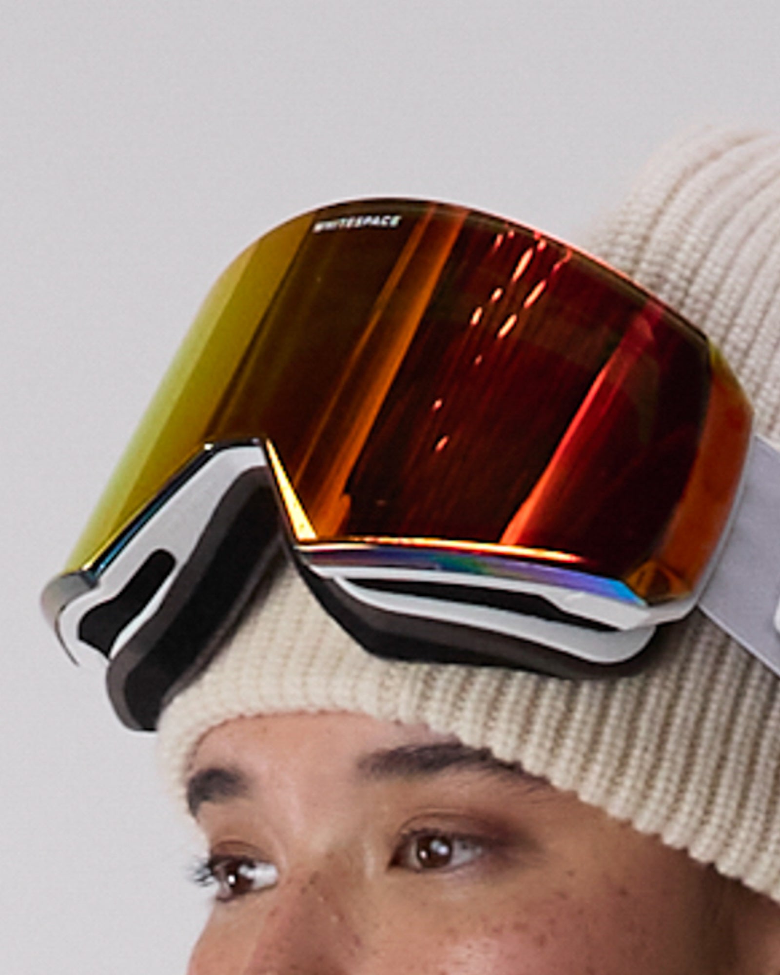 Close-up of a person wearing WHITESPACE MD Icon goggles in white over a cream beanie, on a white background.