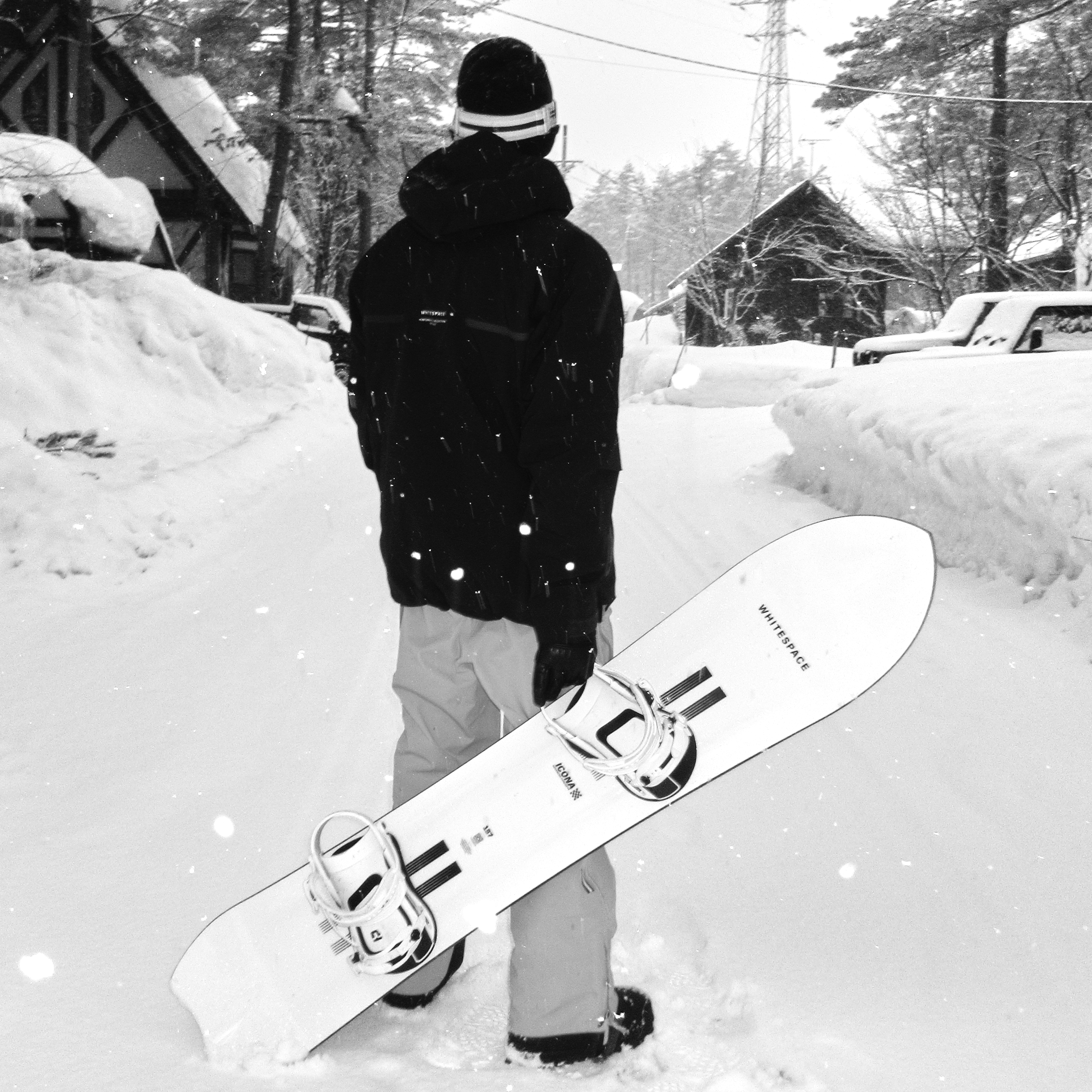 Person holding a snowboard with WHITESPACE branding in a snowy landscape with trees and houses in the background.