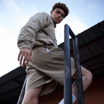 Man, between metal railing with a neutral background, wearing WHITESPACE windbreaker jacket in desert taupe and matching volley shorts.
