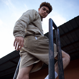 Man, between metal railing with a neutral background, wearing WHITESPACE windbreaker jacket in desert taupe and matching volley shorts.