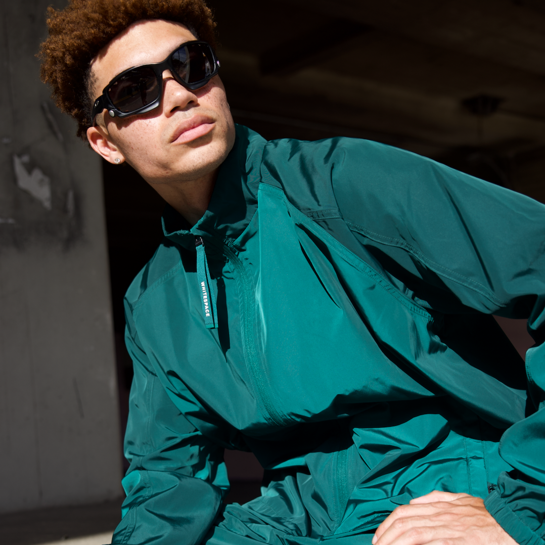 Man, in a seated position, wearing WHITESPACE rainforest green windbreaker jacket, against a dark background.