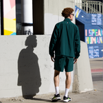 Man, from the back, wearing a WHITESPACE rainforest green windbreaker jacket and volley shorts standing on a sidewalk with a concrete wall and colorful signs in the background.