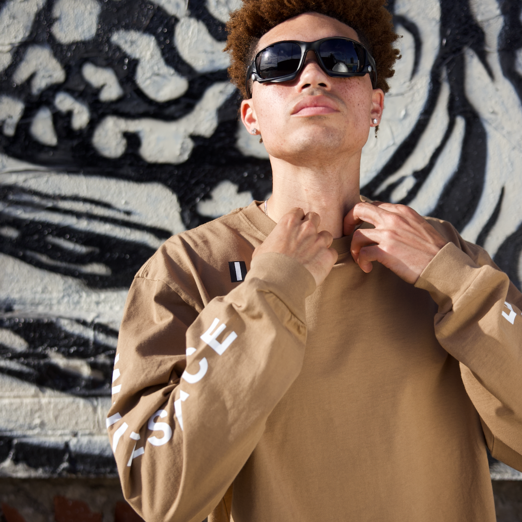 Man wearing a brown WHITESPACE Premium Graphic LS tee, standing against a graffiti-covered wall.
