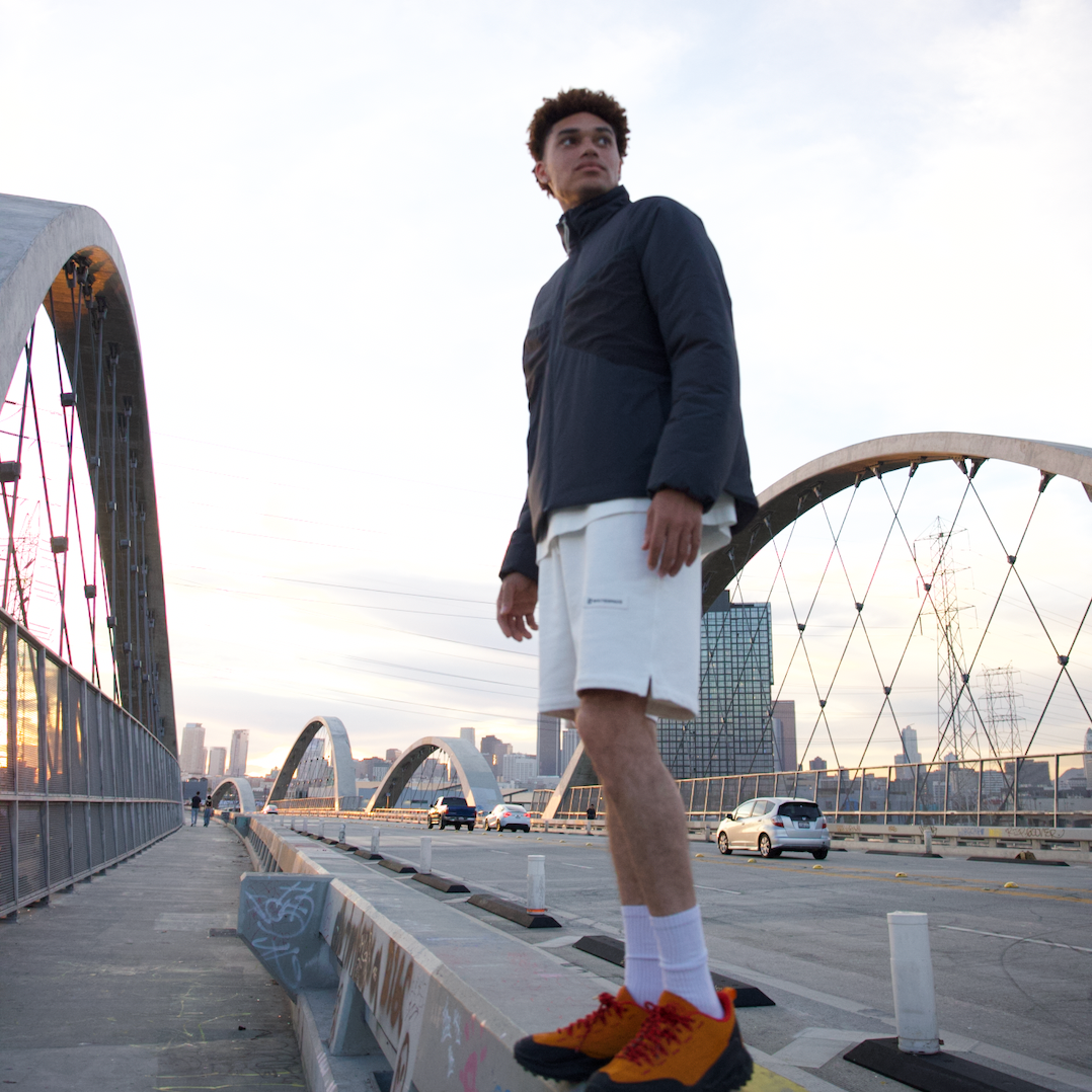 Man standing on a bridge with a cityscape in the background, wearing a WHITESPACE black quarter zip and beige shorts.