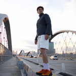 Man standing on a bridge with a cityscape in the background, wearing a WHITESPACE black quarter zip and beige shorts.